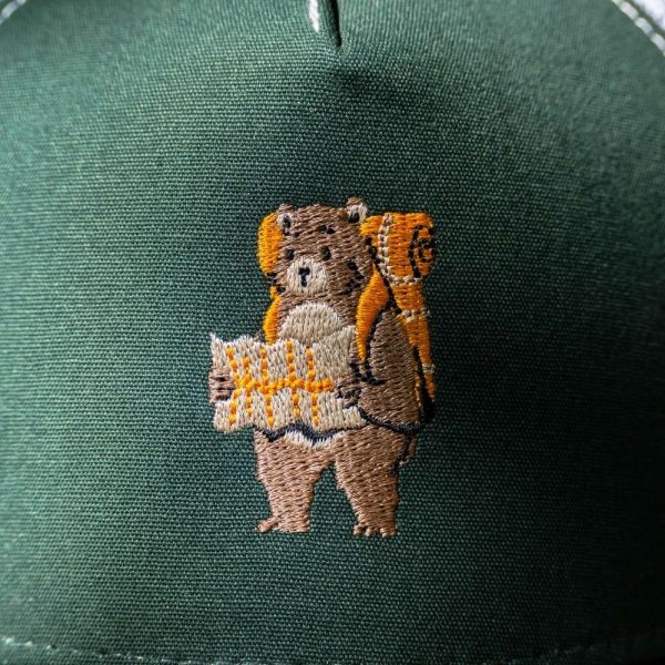 Close-up of an embroidered bear with a backpack and map on a dark green cap.