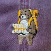Close-up of an embroidered bear with a backpack and map on a faded burgundy cap.