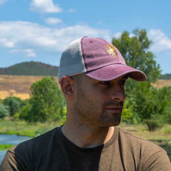 Man wearing a vintage-style faded burgundy trucker cap with embroidered bear carrying a map and backpack, outdoors.