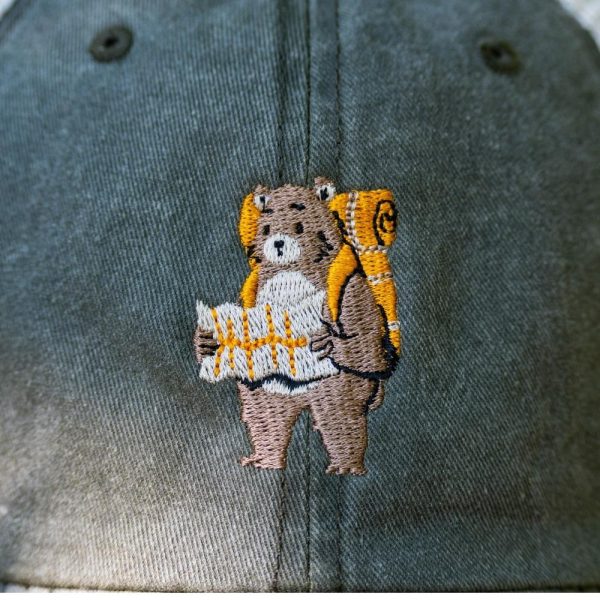 shapka-s-kozirka-meka-mrezha-zelena Close-up of a vintage-style gray cap featuring an embroidered bear with a backpack and map.