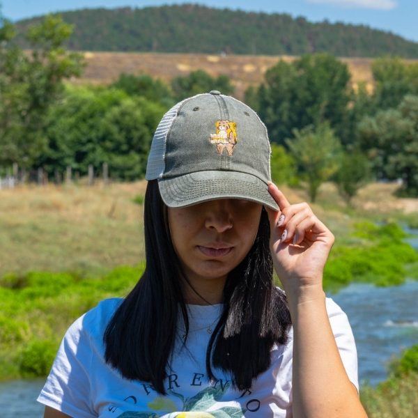 shapka-s-kozirka-meka-mrezha-zelena-zhenska Woman wearing a vintage-style gray cap with an embroidered bear and mesh back, adjusting the brim outdoors.