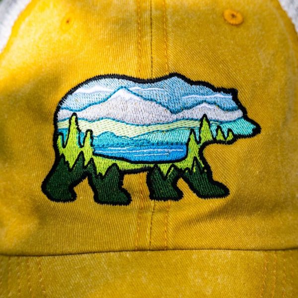 Close-up of embroidered bear patch on a yellow vintage trucker hat, featuring a landscape design with mountains, trees, and a lake.