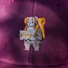 Close-up of an embroidered bear with a backpack and map on a burgundy cap, highlighting the intricate stitching details.