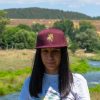 Woman wearing a burgundy snapback cap with an embroidered bear holding a map, standing outdoors with a river and green hills in the background.