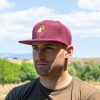 Man wearing a burgundy snapback cap with an embroidered bear holding a map, standing outdoors with a scenic nature background.
