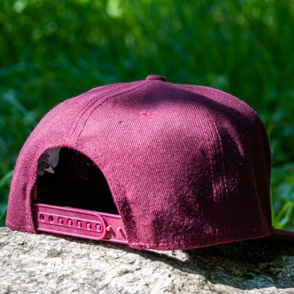 Back view of a burgundy snapback cap with an adjustable strap, placed on a rock with a green nature background.