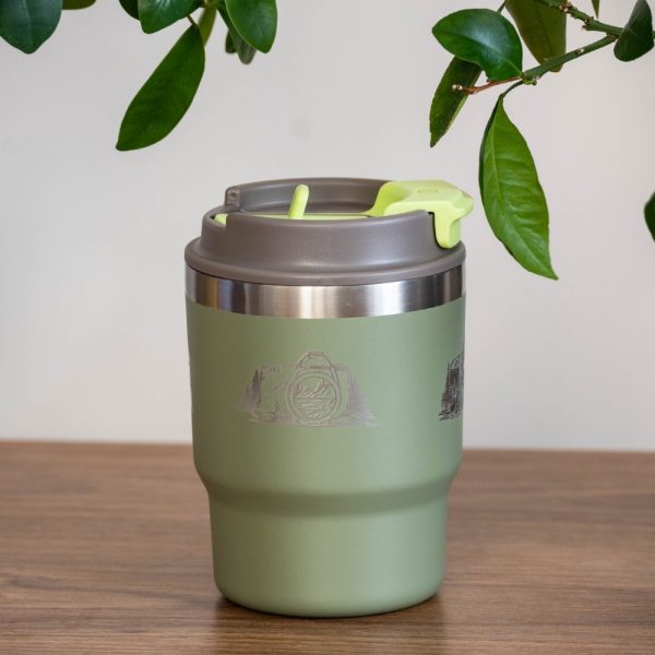 thermo-chasha-za-kafe-chai-photographer Green stainless steel thermal mug (350 ml) with a laser-engraved camera and mountain design. The lid is closed, and green leaves hang above it.