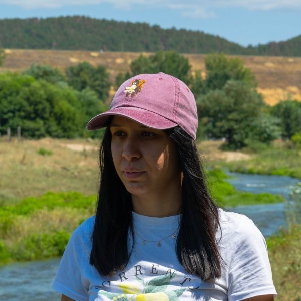 Woman wearing a vintage pink baseball cap with an embroidered bear holding a map, standing in a scenic outdoor location.