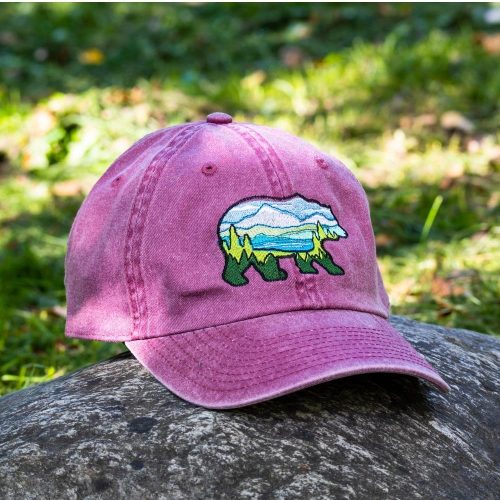 vintage-shapka-s-kozirka-bordo-emroidery Vintage pink baseball cap with an embroidered bear silhouette filled with a scenic mountain and forest design, placed on a rock outdoors.