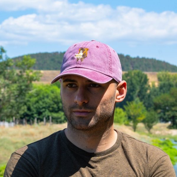 Man wearing a vintage pink baseball cap with an embroidered bear holding a map, standing outdoors in nature.