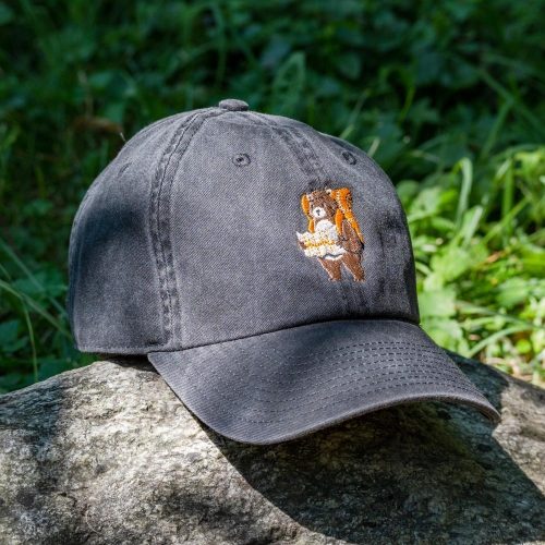 vintage-shapka-s-kozirka-cherna Vintage black baseball cap with an embroidered bear holding a map, placed on a rock with a green forest background.