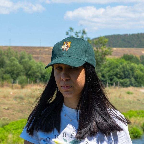 Woman wearing a dark green baseball cap with an embroidered bear holding a map, standing outdoors with hills and trees in the background.