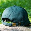 Back view of a dark green baseball cap with an adjustable brass buckle strap, placed on a rock with a blurred green nature background.