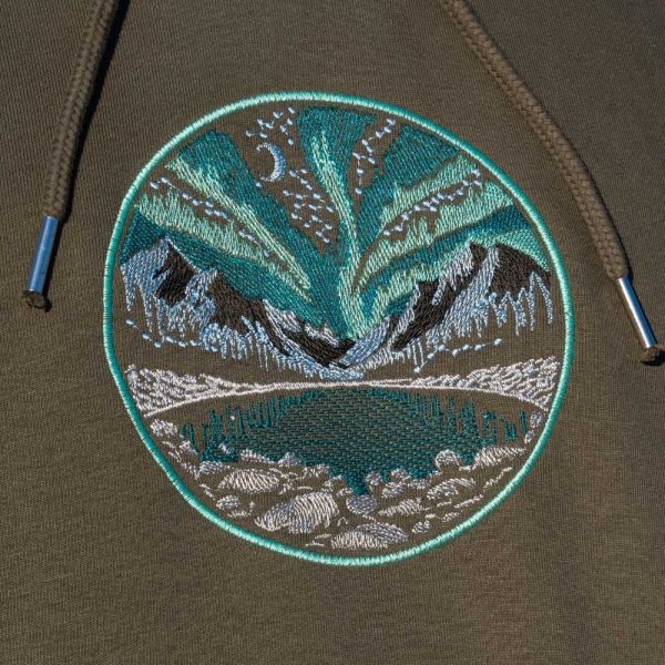 Close-up of an embroidered Northern Lights design on an olive green hoodie, featuring mountains, a lake, and a night sky.