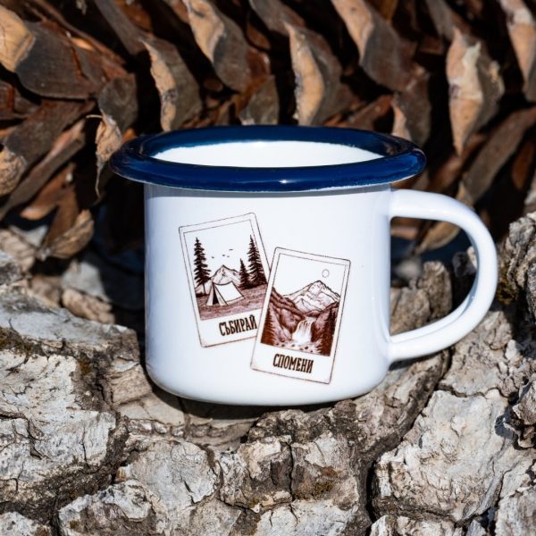 100ml - malko- kanche - 3oz White enamel mug with blue rim and illustration of mountains, tent and the text “Collect Memories”, placed outdoors on bark.