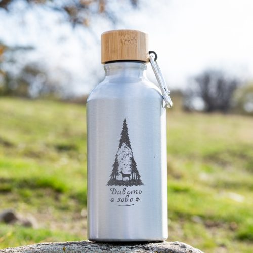 butilka-metalna-za-kafe Stainless steel water bottle with bamboo cap and engraved mountain-and-deer design, standing outdoors on a rock.