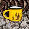 Yellow enamel mug with black rim and illustration of a campfire, tent, and pine trees with the text “Don’t burn the forest, light the fire in your soul”, photographed outdoors on bark.
