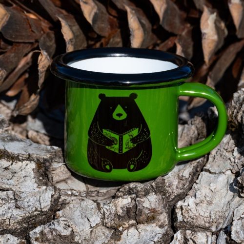 kanche-zeleno-s-mechka Green enamel mug with black bear reading a book illustration, placed outdoors on bark.