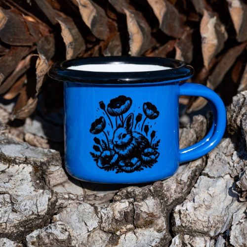 malko-sinio-kanche Small blue enamel mug with black bunny and poppy flower illustration, placed outdoors on bark.