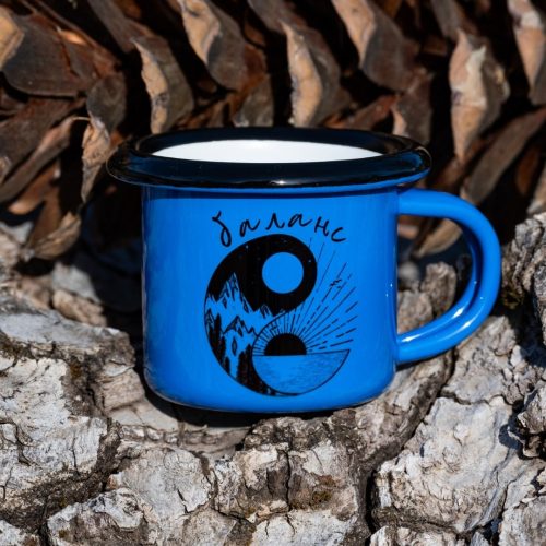 malko-sinio-kanche-balance Small blue enamel mug with yin-yang mountain and sun illustration and text “Balance”, placed outdoors on bark.
