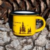 Yellow enamel mug with black rim and illustration of pine trees and a tent, with outdoor adventure design, placed on bark.