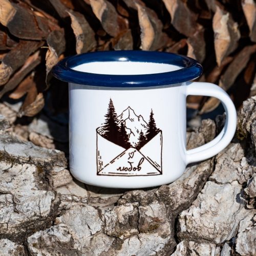 mini-malko-kanche White enamel mug with blue rim and illustration of envelope with trees and mountain inside, and the text “Love”, placed outdoors on bark.