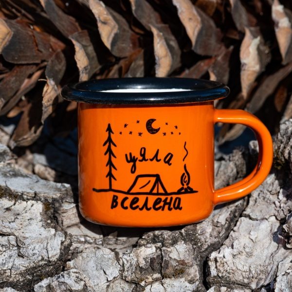 podarak-za-planinar-kanche Small orange enamel mug with camping illustration, tent, and text “My Universe”, placed outdoors on bark.