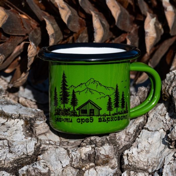 zeleno-kanche-malko Green enamel mug with black illustration of a mountain cabin, pine trees and the text “Live Among the Peaks”, placed outdoors on bark.
